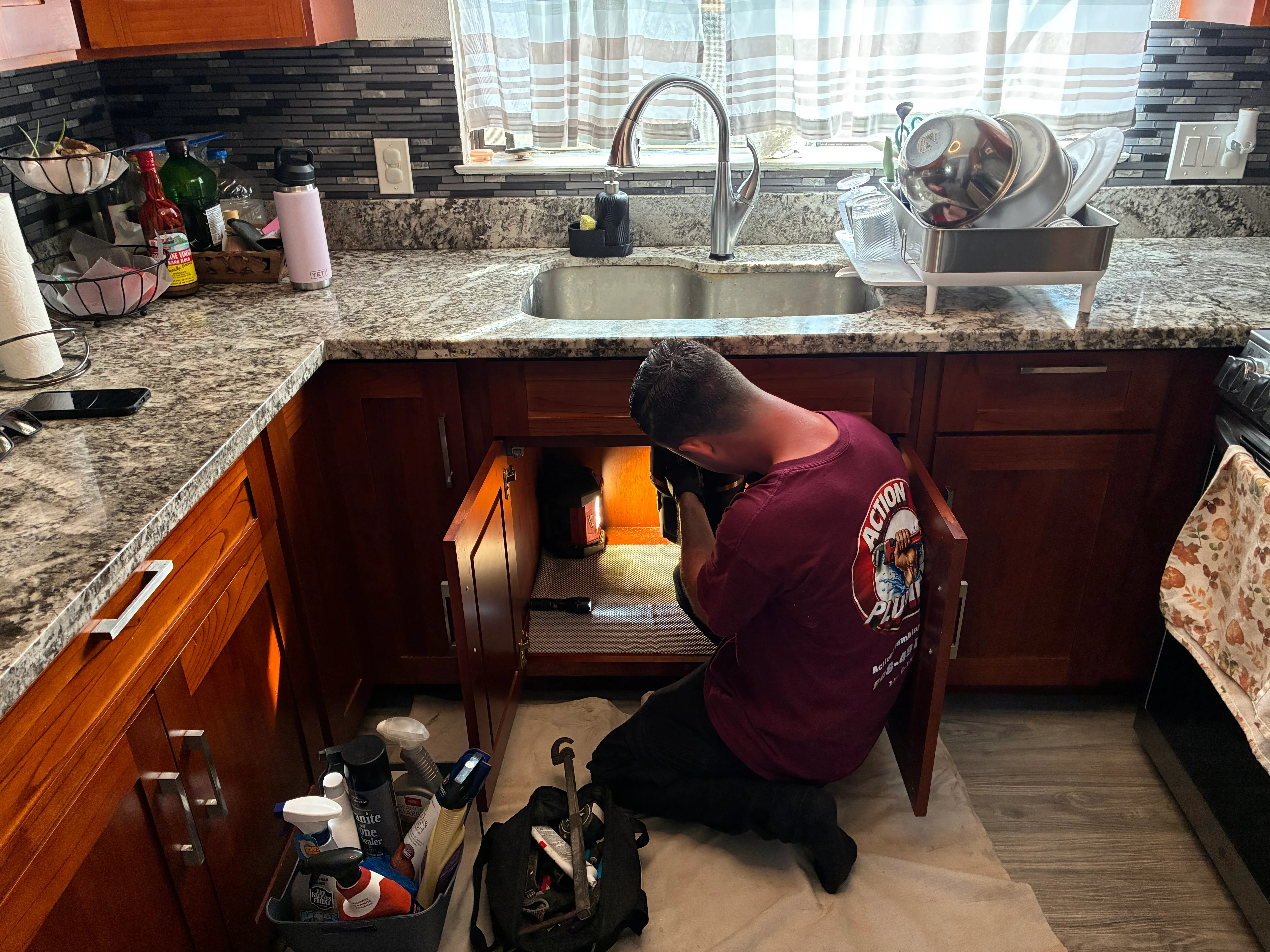 Plumber working under kitchen sink with granite countertop and dark wood cabinets